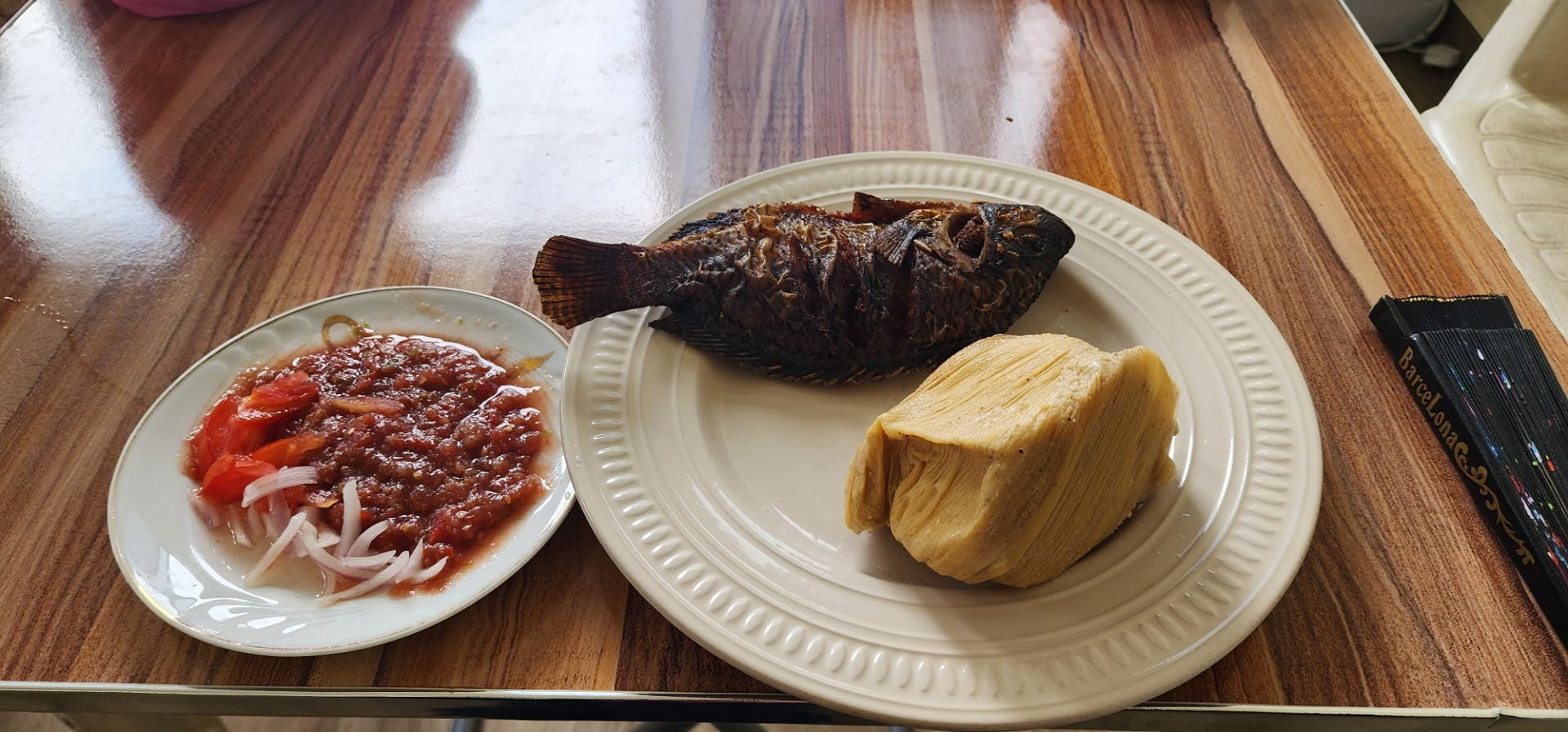 A large plate containing kenkey and a fried tilapia. To the left, a smaller plate containing tomato sauce, onions, and peppers.