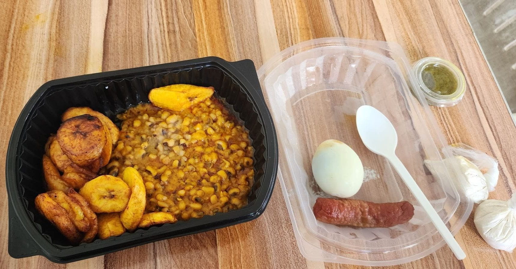 A takeout box containing black eyed peas and fried plantains. The lid is to the right containing a hard-boiled egg and a chicken sausage.