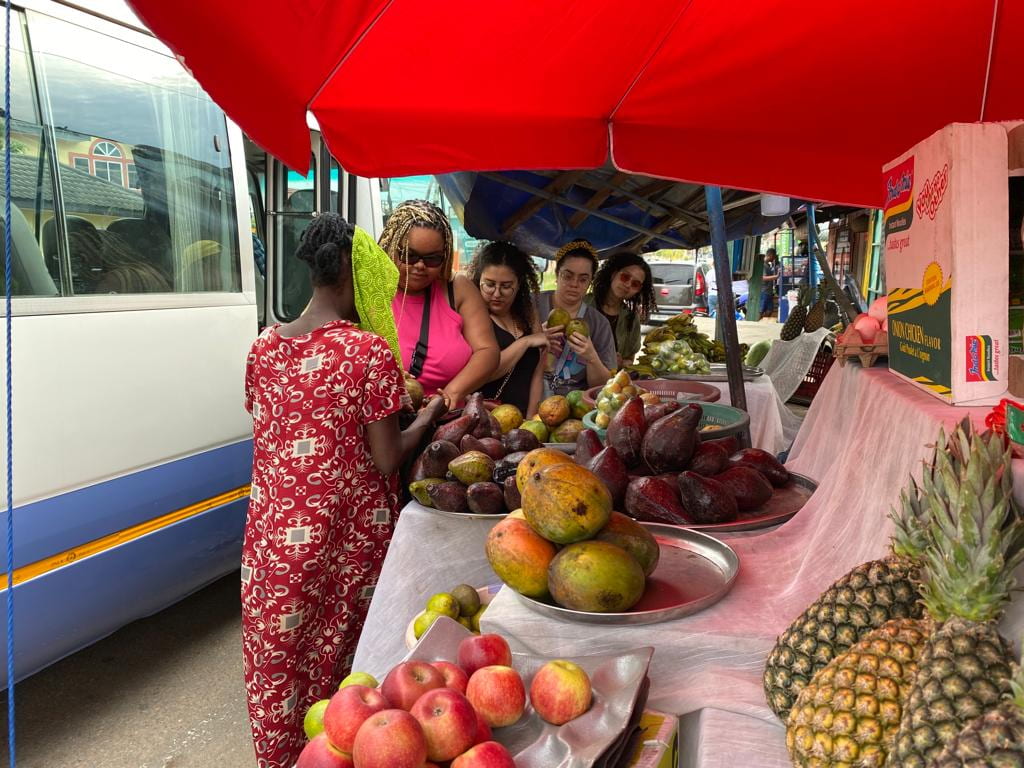 A group of women stand between a bus and a fruit stand, buying mangoes on the side of the street. 